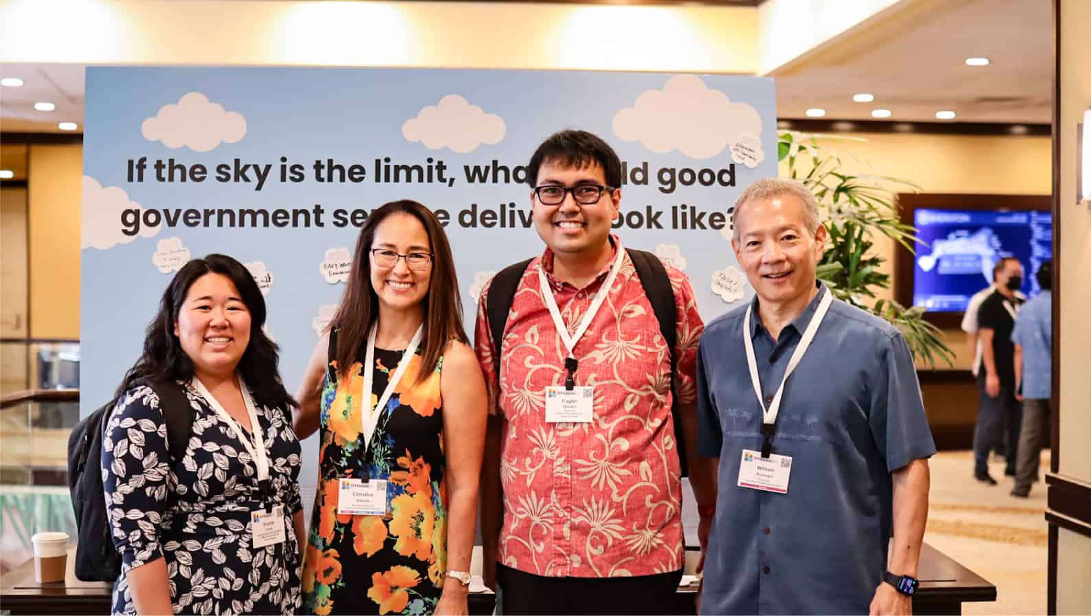 Four people are standing together at a conference, smiling at the camera. They are all wearing conference name tags. Behind them is a colorful backdrop with the text, "If the sky is the limit, what would good government service delivery look like?.