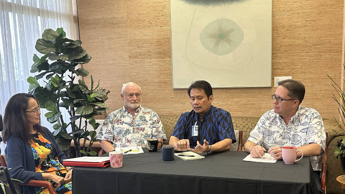 Four people sit around a table covered in a black tablecloth engaging in discussion. Three of them are men wearing patterned shirts, and one is a woman in a floral dress. The setting appears to be a well-lit room with a plant and abstract art in the background.