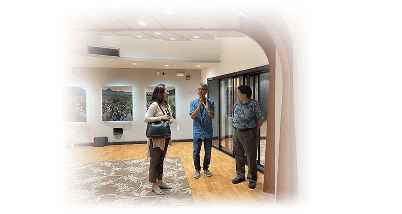 Three individuals are standing in a modern interior space with wooden ceiling arches. Wall-mounted artworks depicting nature scenes hang in the background. Recessed lighting illuminates the room, which features a patterned carpet and wooden flooring.