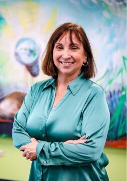 A woman with shoulder-length brown hair is standing with her arms crossed, smiling, and wearing a teal blouse. The background features a colorful mural depicting natural elements with a green and blue color scheme.