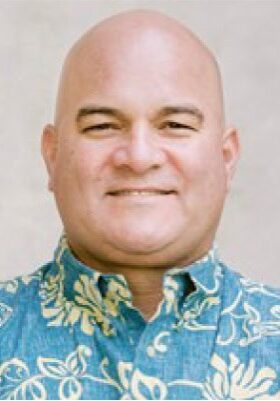 A bald man with a light complexion is smiling at the camera. He is wearing a blue shirt with a floral pattern. The background is plain and beige.