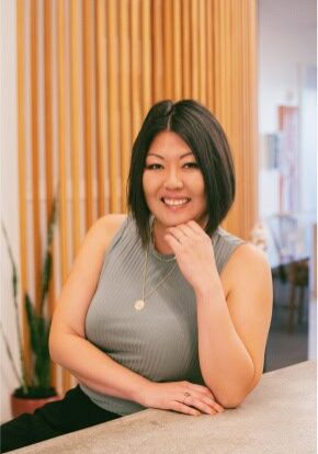 A woman with shoulder-length black hair is smiling and resting her chin on her right hand. She is wearing a gray sleeveless top and a gold necklace with a circular pendant. She is standing in front of a wooden panel backdrop with a plant visible in the background.