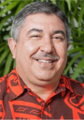 A smiling man with short, graying hair, a moustache, and wearing a red patterned shirt stands in front of a lush green backdrop.