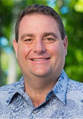 A person with short brown hair and a cheerful smile is standing outdoors with a green, leafy background. They are wearing a blue patterned shirt.