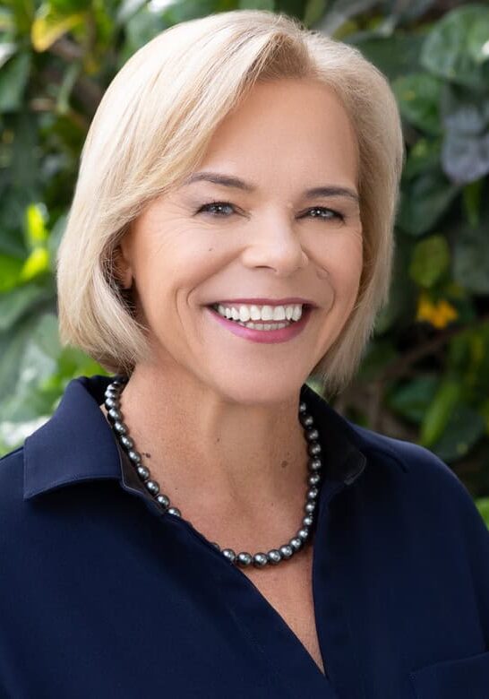 A smiling woman with short blonde hair is wearing a dark blouse and pearl necklace. She is standing in front of a background of green foliage.