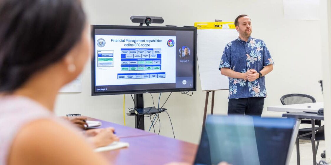 A person in a floral shirt stands beside a display screen giving a presentation to a small group in a conference room. The screen shows a financial management flowchart. Other attendees are seated, one with a laptop, focusing on the presentation.